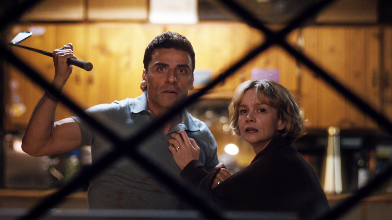 "A man holding a golf club and a woman stand close together looking alarmed inside a dimly lit kitchen, viewed through a window with a crisscross frame."